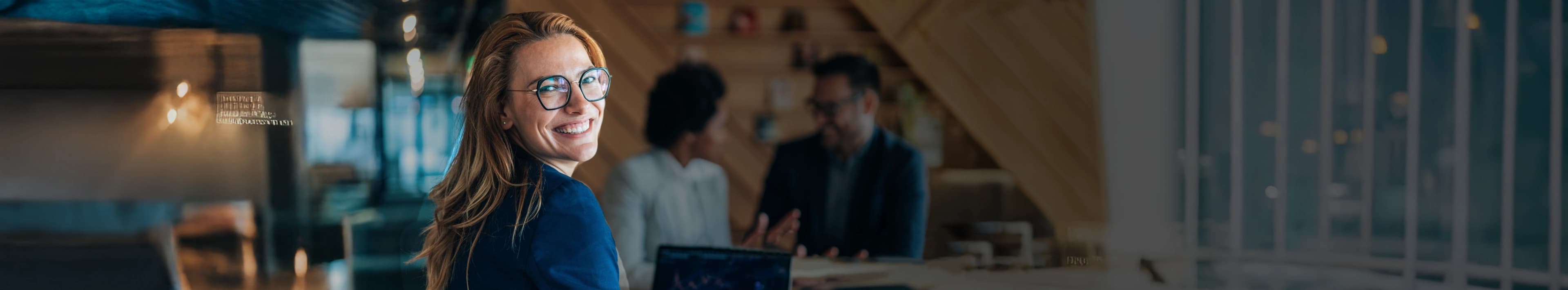 A businesswoman with glasses smiles, with her team working in the blurred background.
