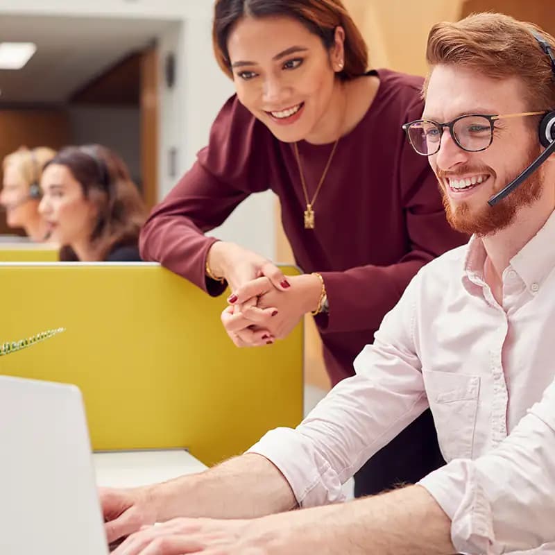 A male and female support agent smiling while they assist a customer on a computer in a call center.