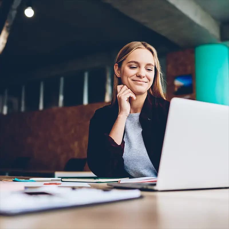 Eine lächelnde Freelancerin schaut während der Arbeit in einem Café auf ihren Laptop.