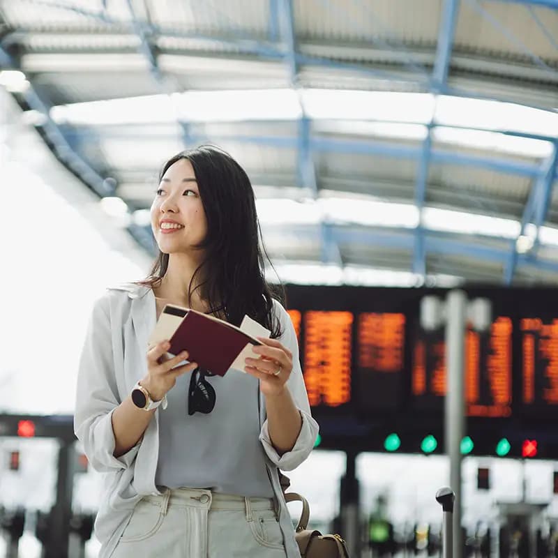 A young, happy woman stands at an airport holding a passport and a ticket.