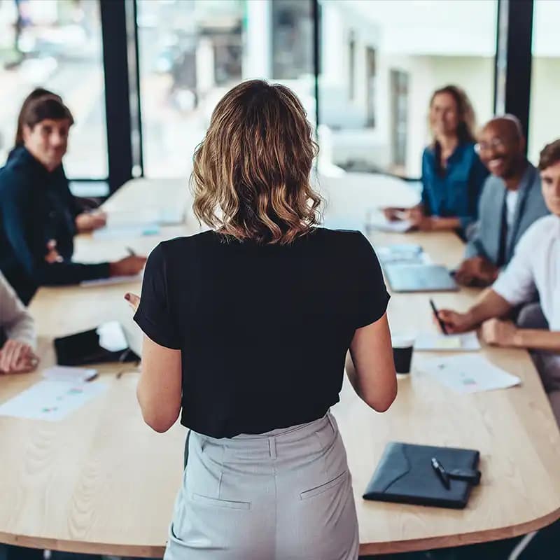 A businesswoman stands and presents ideas to her team seated around a large table during a meeting.