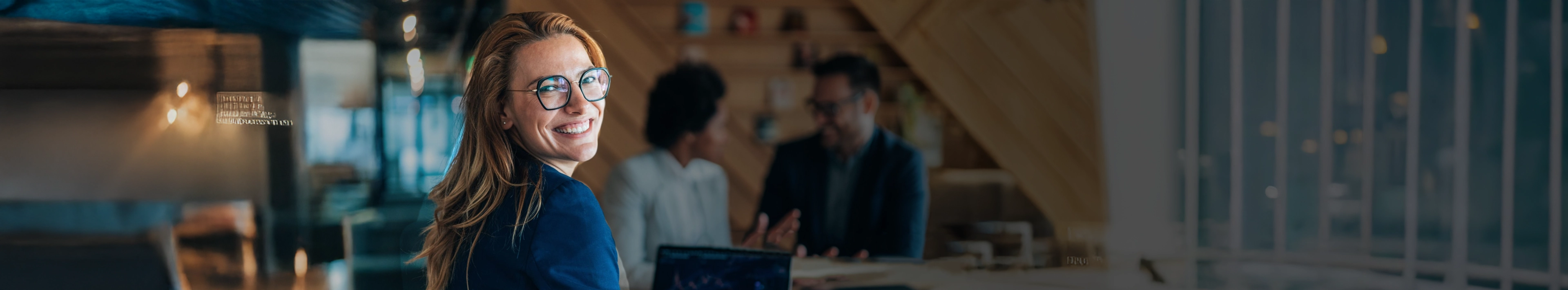 A businesswoman with glasses smiles, with her team working in the blurred background.